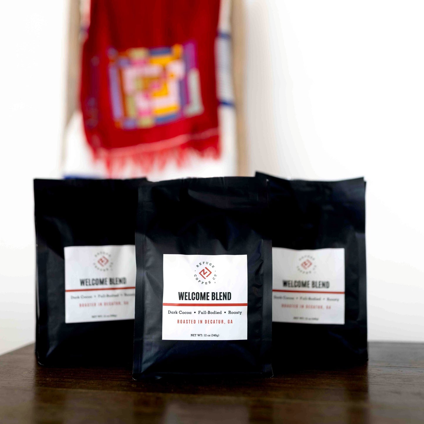 Three black coffee bags labeled 'Welcome Blend' on a wooden surface with a blurred colorful fabric in the background.